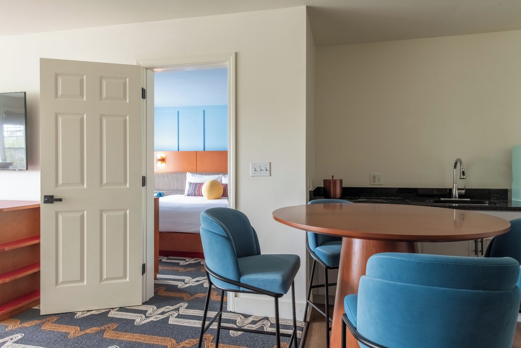 Hotelkamer met een bed, een ronde houten tafel met blauwe stoelen, een kitchenette en een deur naar een andere kamer.