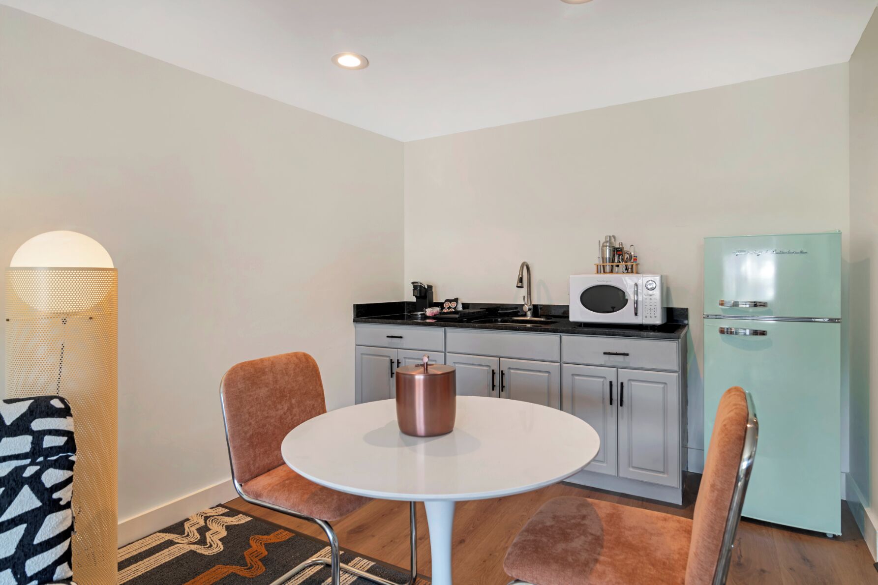 A compact kitchen with a white refrigerator, a microwave, and a coffee maker on the counter. There is a round table with two chairs and a small rug on the floor.
