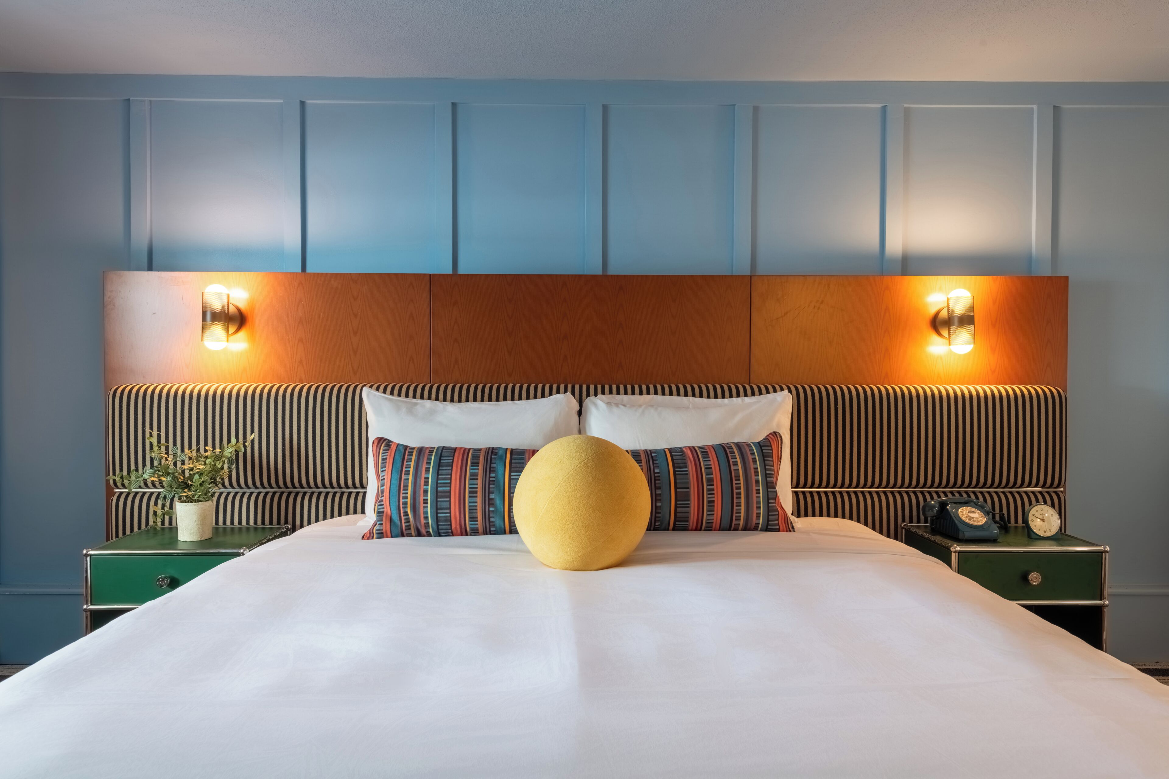 A neatly made bed with a large round pillow, striped pillows, and a headboard with wall-mounted lights.