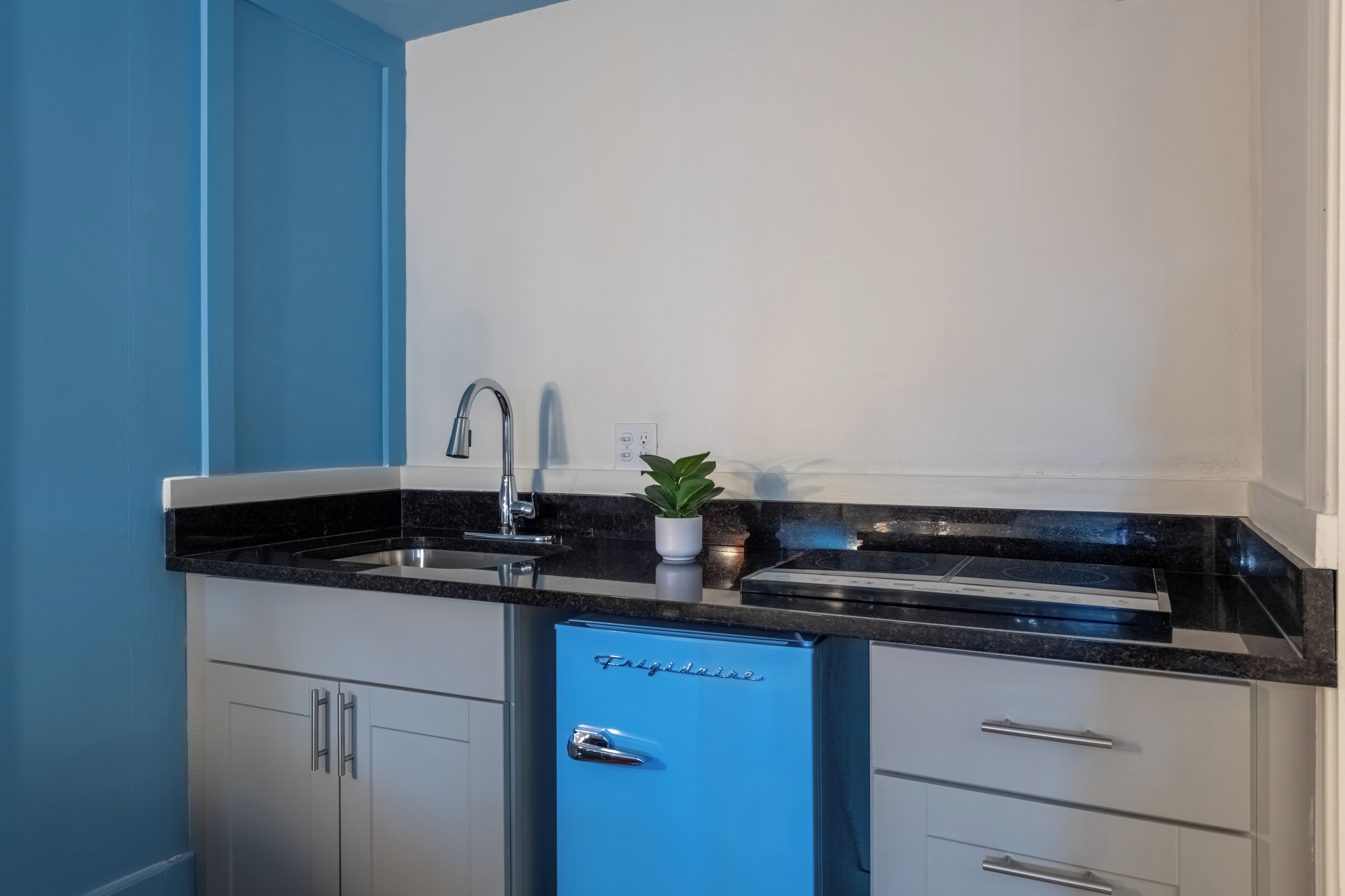 Una cocina compacta con un refrigerador azul, armarios de acero inoxidable, una encimera negra y una pequeña planta en maceta.