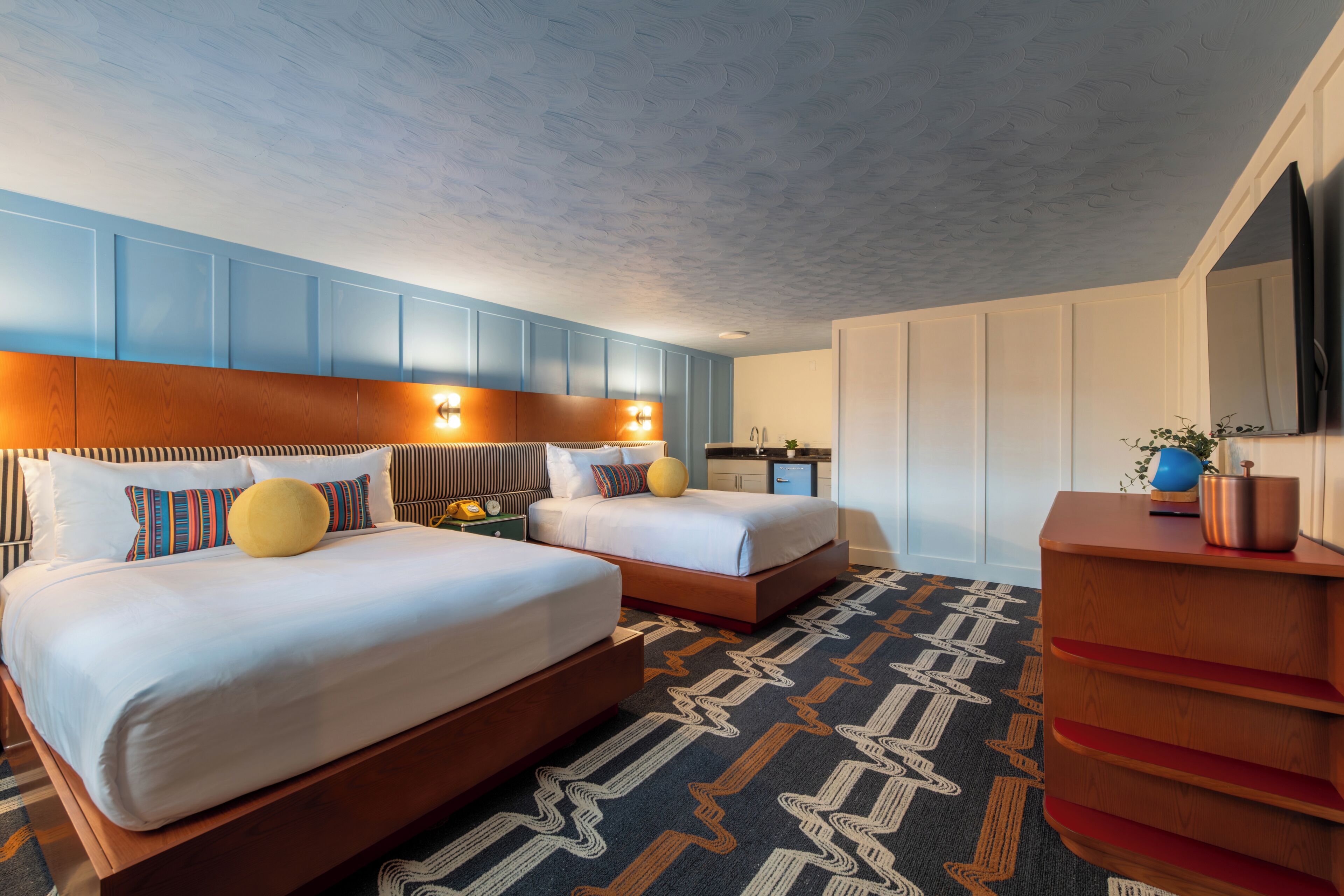 A hotel room with two beds, a wooden dresser, a TV, and a patterned carpet.