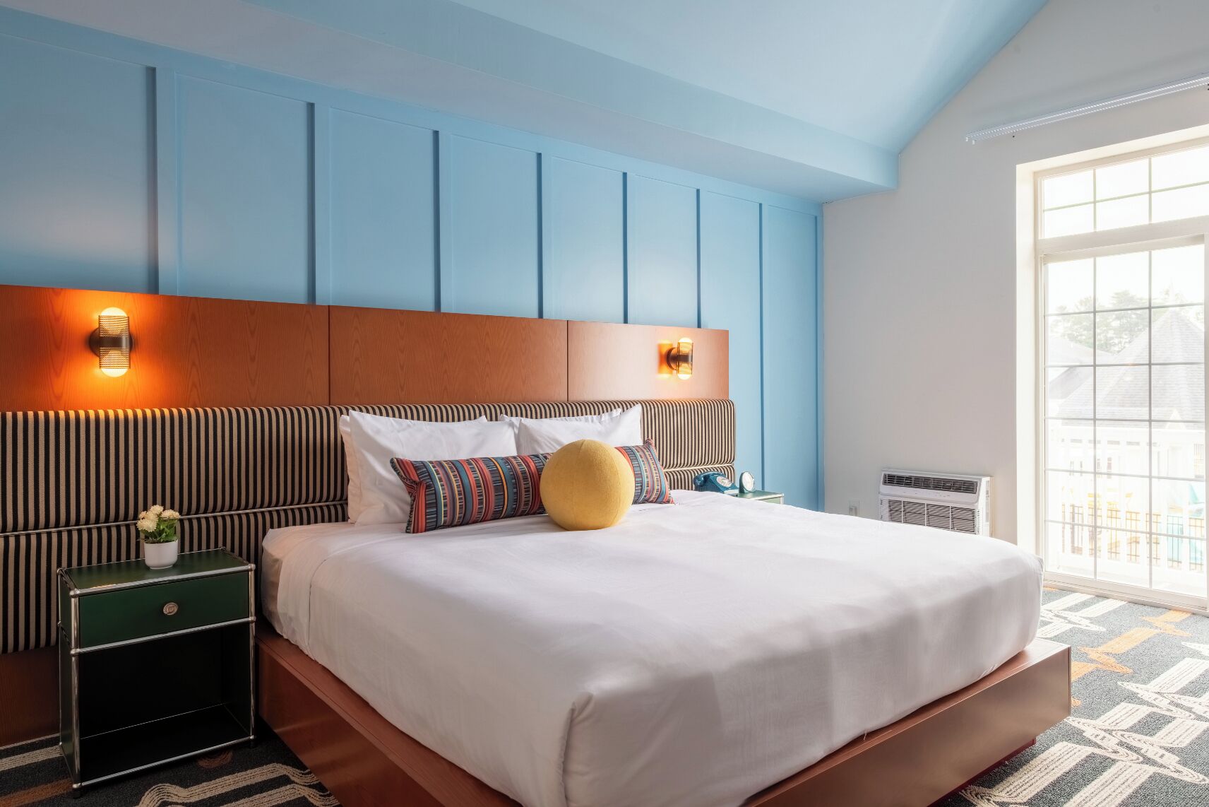 Quarto de hotel com uma cama grande, cabeceira de madeira, mesa de cabeceira e uma mesinha verde, além de uma janela com vista.