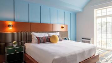 Quarto de hotel com uma cama grande, cabeceira de madeira, mesa de cabeceira e uma mesinha verde, além de uma janela com vista.
