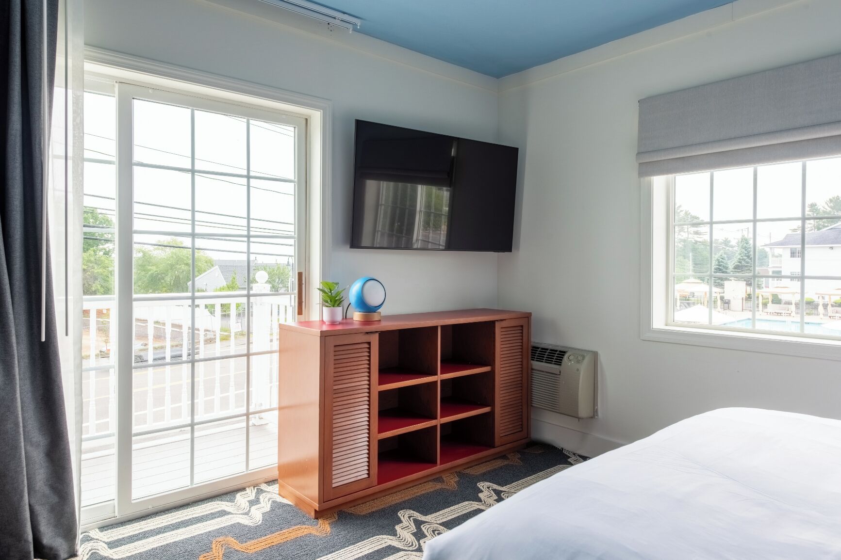 Une chambre d’hôtel avec un lit, un téléviseur fixé au mur, une armoire en bois avec des étagères, un radiateur et de grandes fenêtres donnant sur l’extérieur.