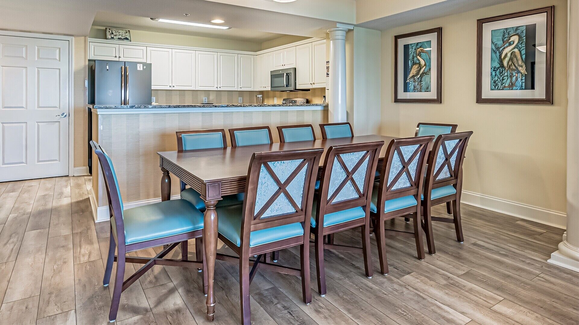 A dining area with a wooden table and blue upholstered chairs, a kitchen with white cabinets, and two framed wall art pieces.