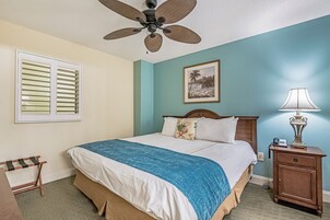 Un dormitorio con una cama, ventilador de techo, mesita de noche con lámpara, un cuadro en la pared y una ventana con persianas.