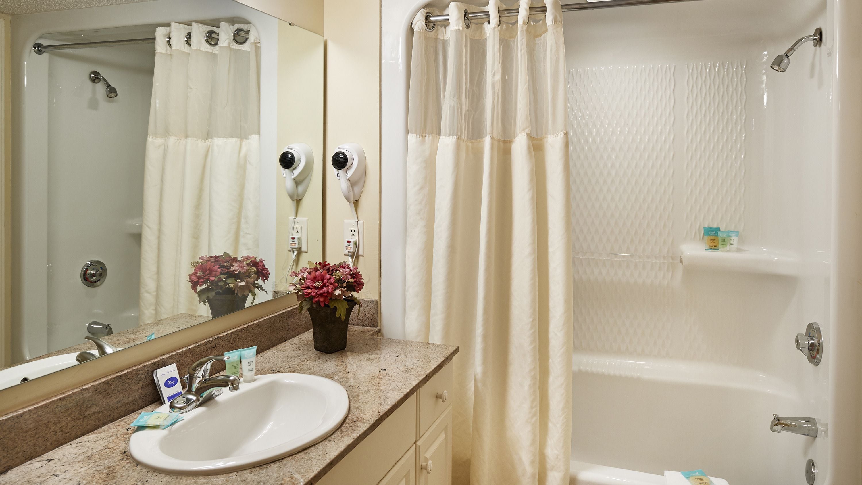 A bathroom with a sink, mirror, shower, and two hair dryers on the wall.