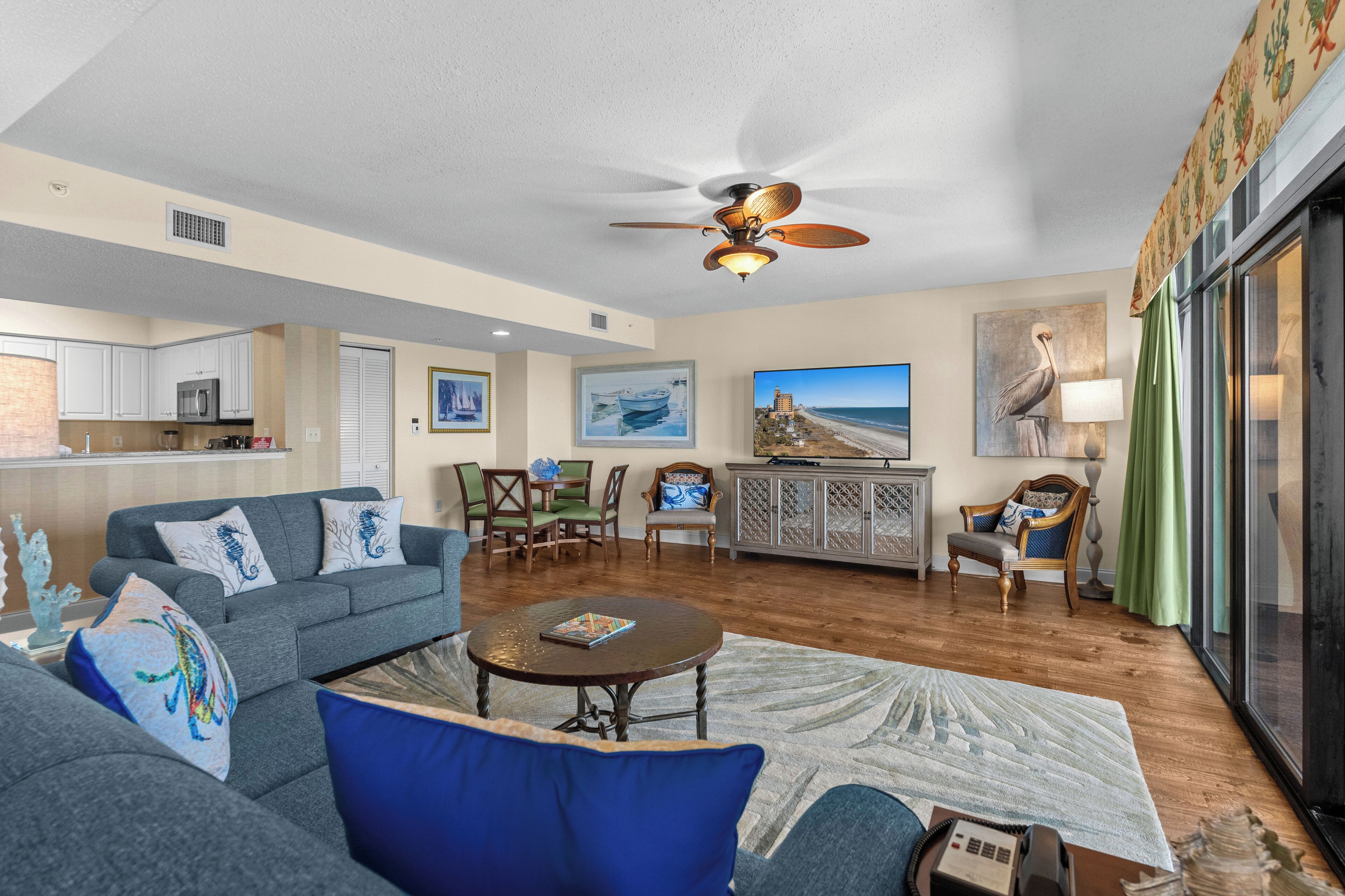 A spacious living area with a grey sofa, a round coffee table, and a ceiling fan. There is a kitchen area with white cabinets and a microwave. A large window with a view of the beach is visible.