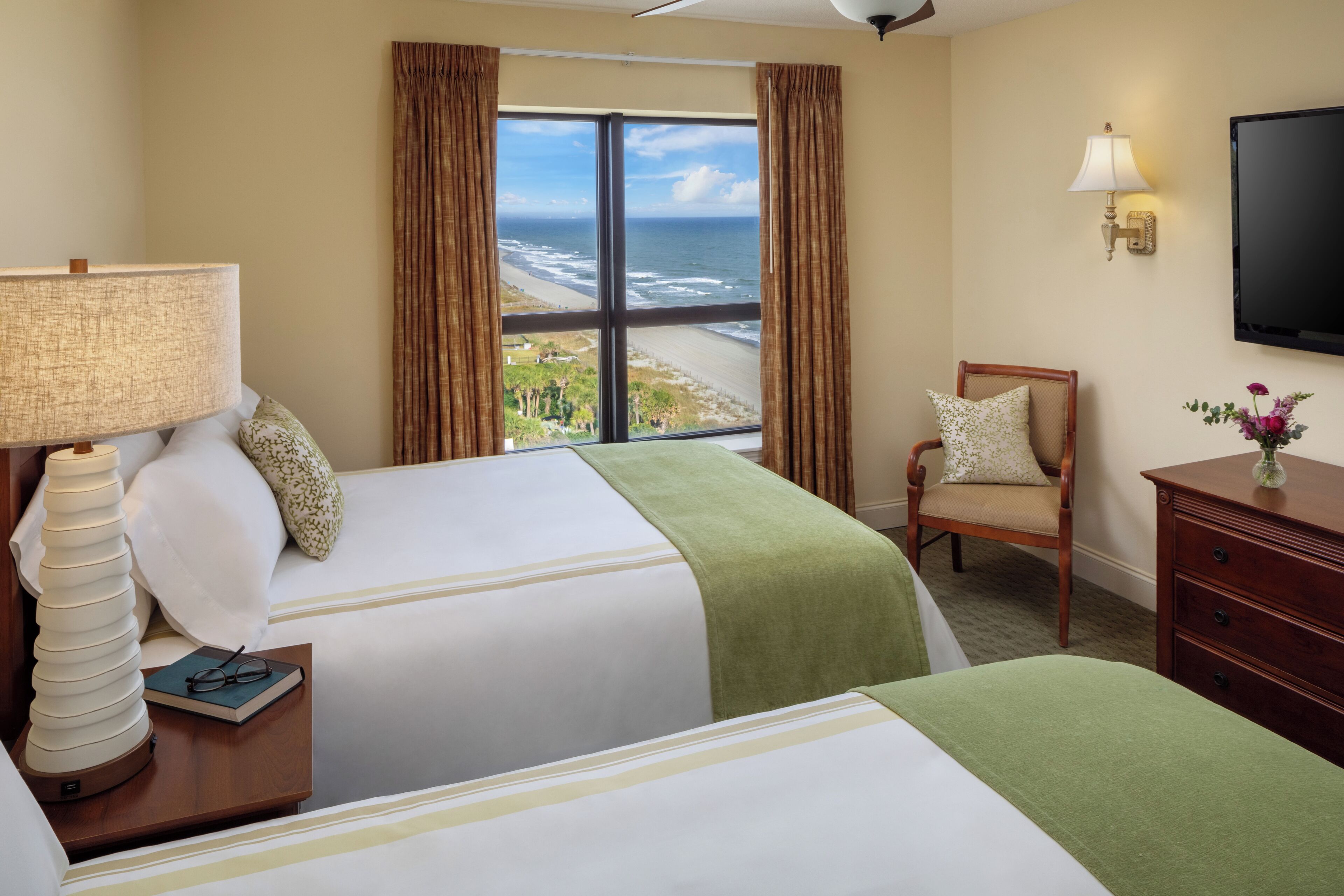 A hotel room with two beds, a view of the ocean, a television, and a lamp.