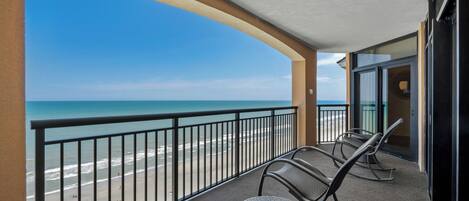 A balcony with a view of the beach and ocean. There are two wicker chairs and a small round table.