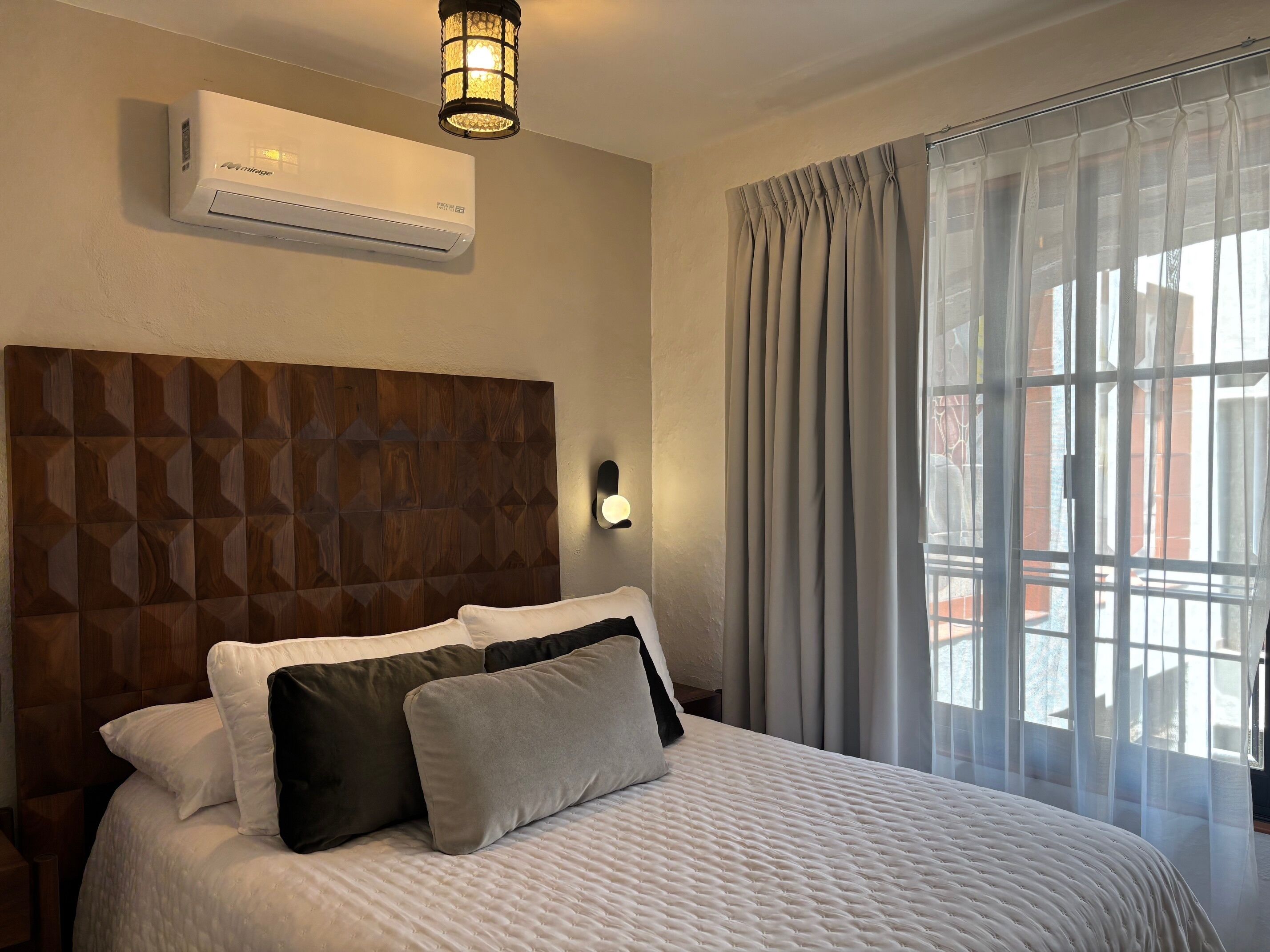 Une chambre d’hôtel avec un lit, un climatiseur fixé au mur, une tête de lit à motifs géométriques, une lampe de chevet et une fenêtre avec des rideaux.