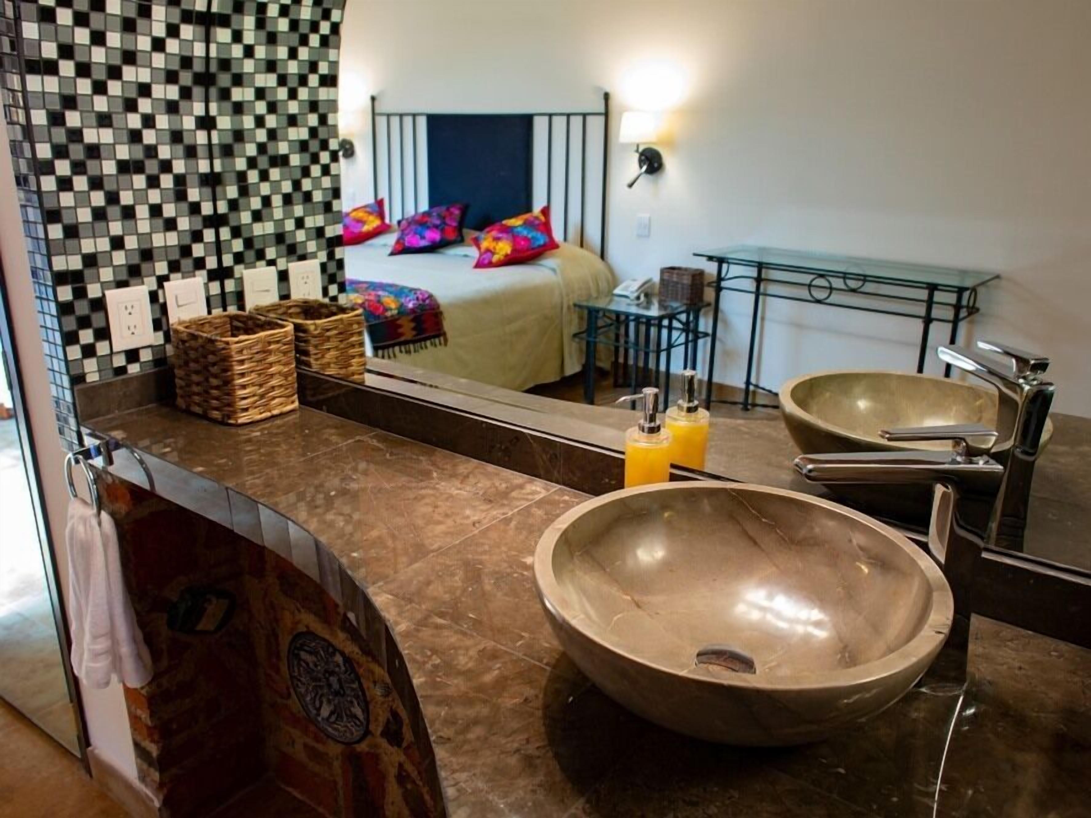 Une salle de bain avec un grand miroir, deux lavabos et un lit visible dans le reflet.