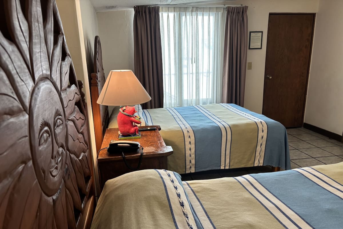 Hotellihuone, jossa on kaksi sänkyä, puinen pääty, yöpöytä lampun ja puhelimen kanssa sekä ikkuna, jossa on verhot.