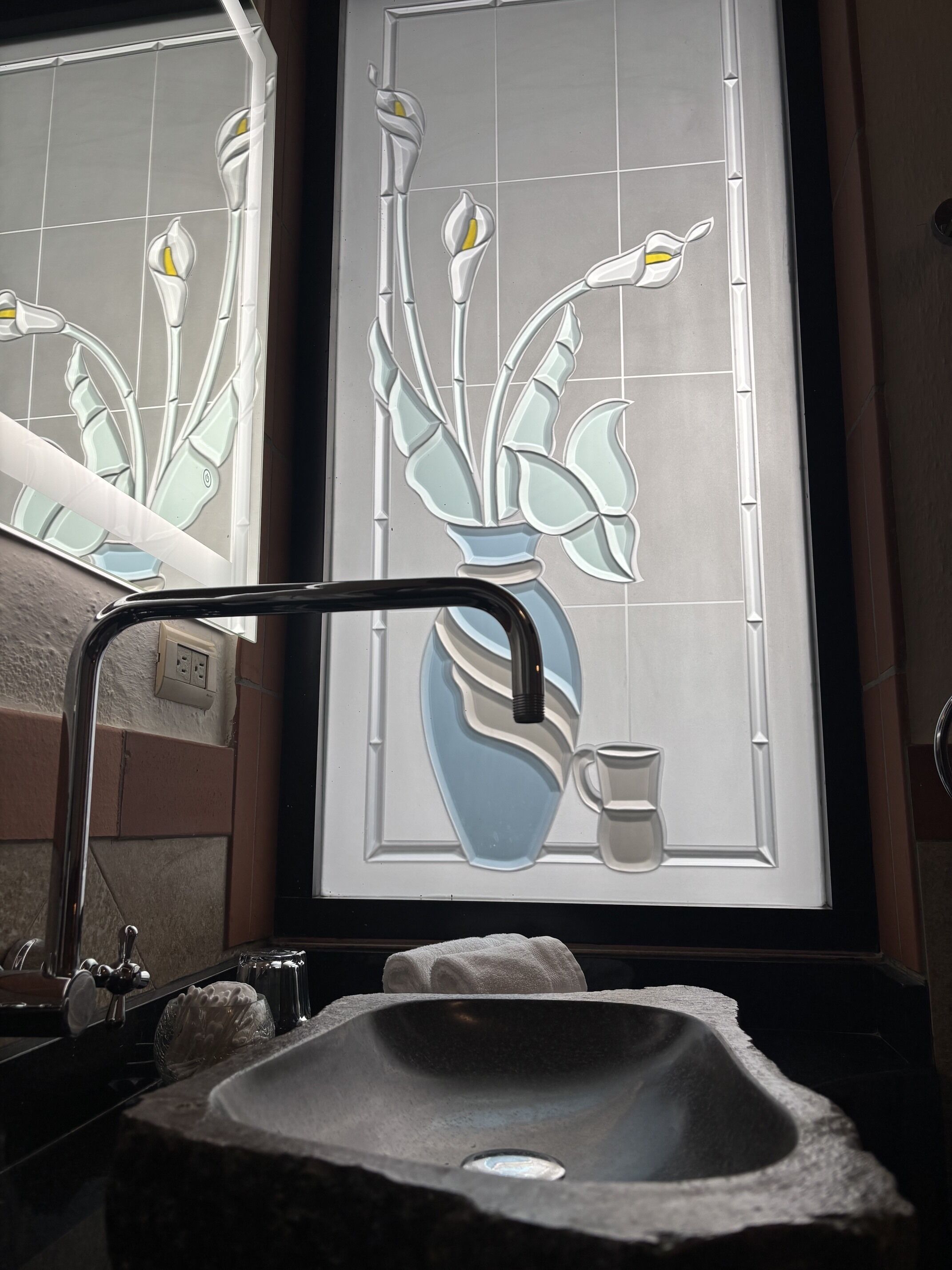 A bathroom with a stone sink, a mirror, and a stained glass window depicting flowers and a cup.