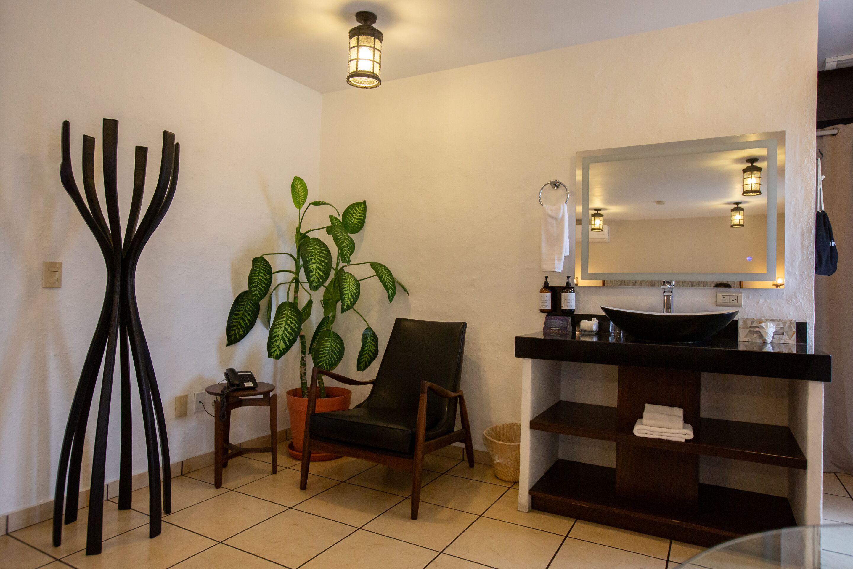 Un baño con lavamanos negro, un espejo, una silla, una planta en maceta y una estructura decorativa negra.