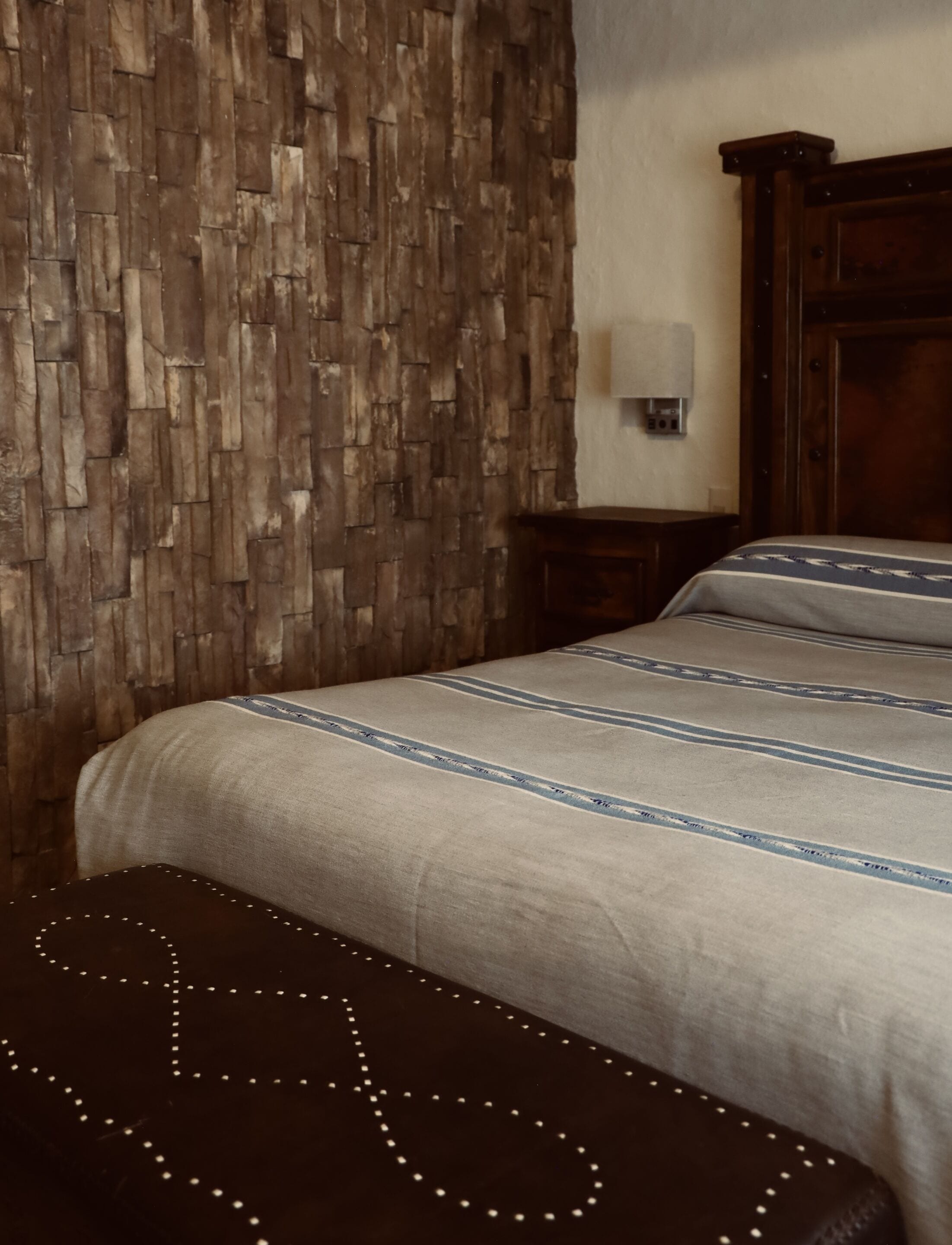 A bedroom with a stone-patterned wall, a wooden headboard, a bed with striped bedding, and a bench with lights.