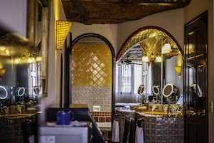 Un baño con un espejo grande, un lavabo y una tocador. Hay varios espejos y un ventilador de techo. Las paredes tienen azulejos con diseño.