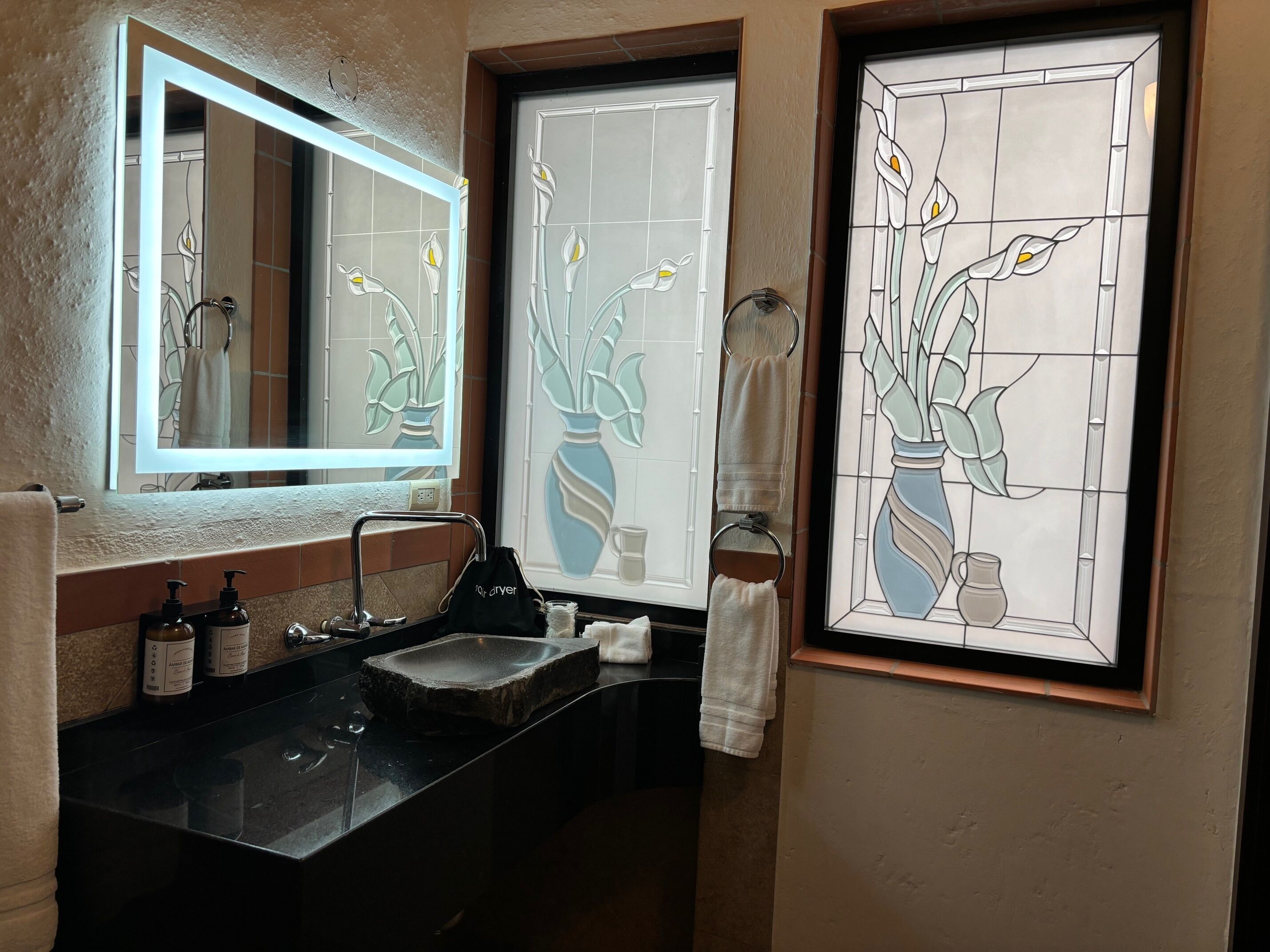 Un baño con un lavabo negro, dos botellas de jabón para manos, un perchero y un espejo.