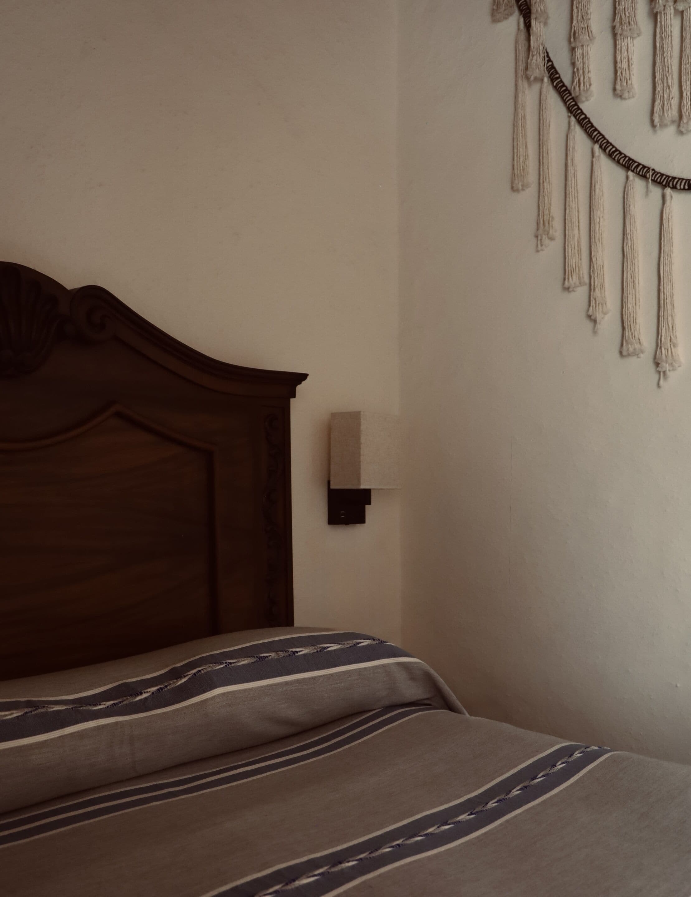 Una cama con cabecera de madera, una lámpara de pared y un tapiz de macramé.