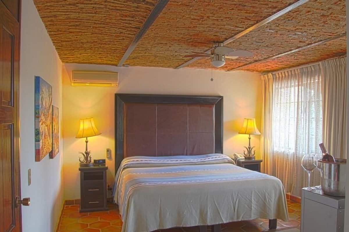 Hotellihuone, jossa on suuri sänky, kaksi yöpöytää valaisimineen, kattotuuletin, ikkuna verhoineen ja laattalattia.