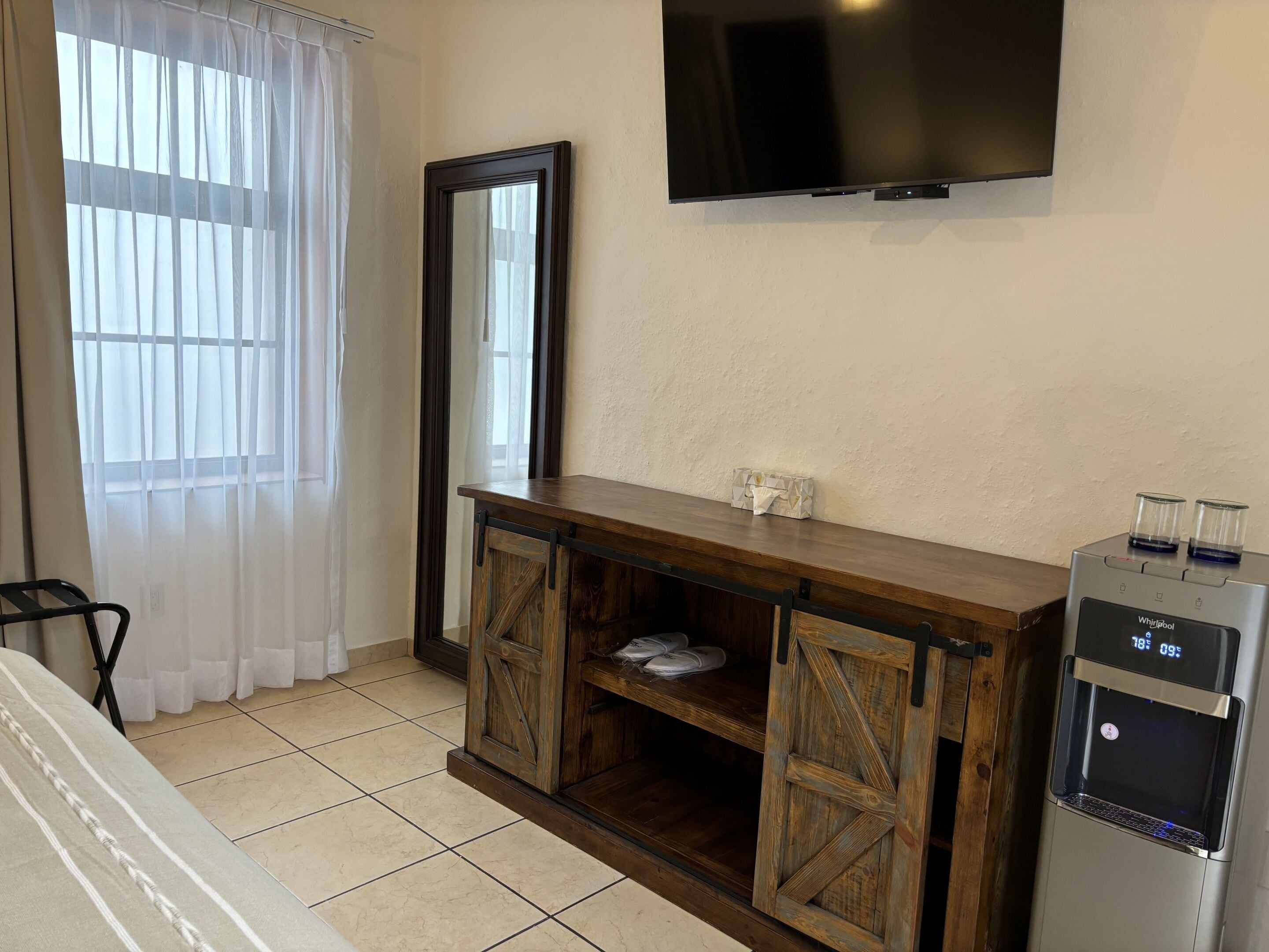Habitació d'hotel amb una taula de console de fusta, un gran mirall, un televisor de pantalla plana penjat a la paret i un dispensador d'aigua.