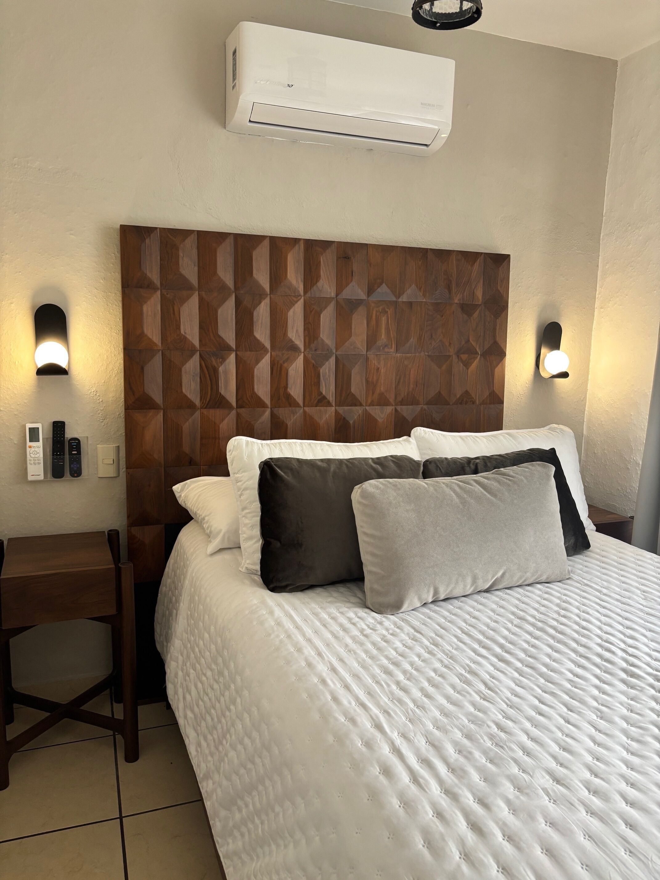 Una habitación de hotel con cabecera de madera, cama con ropa de cama blanca, dos mesitas de noche y aire acondicionado de pared.