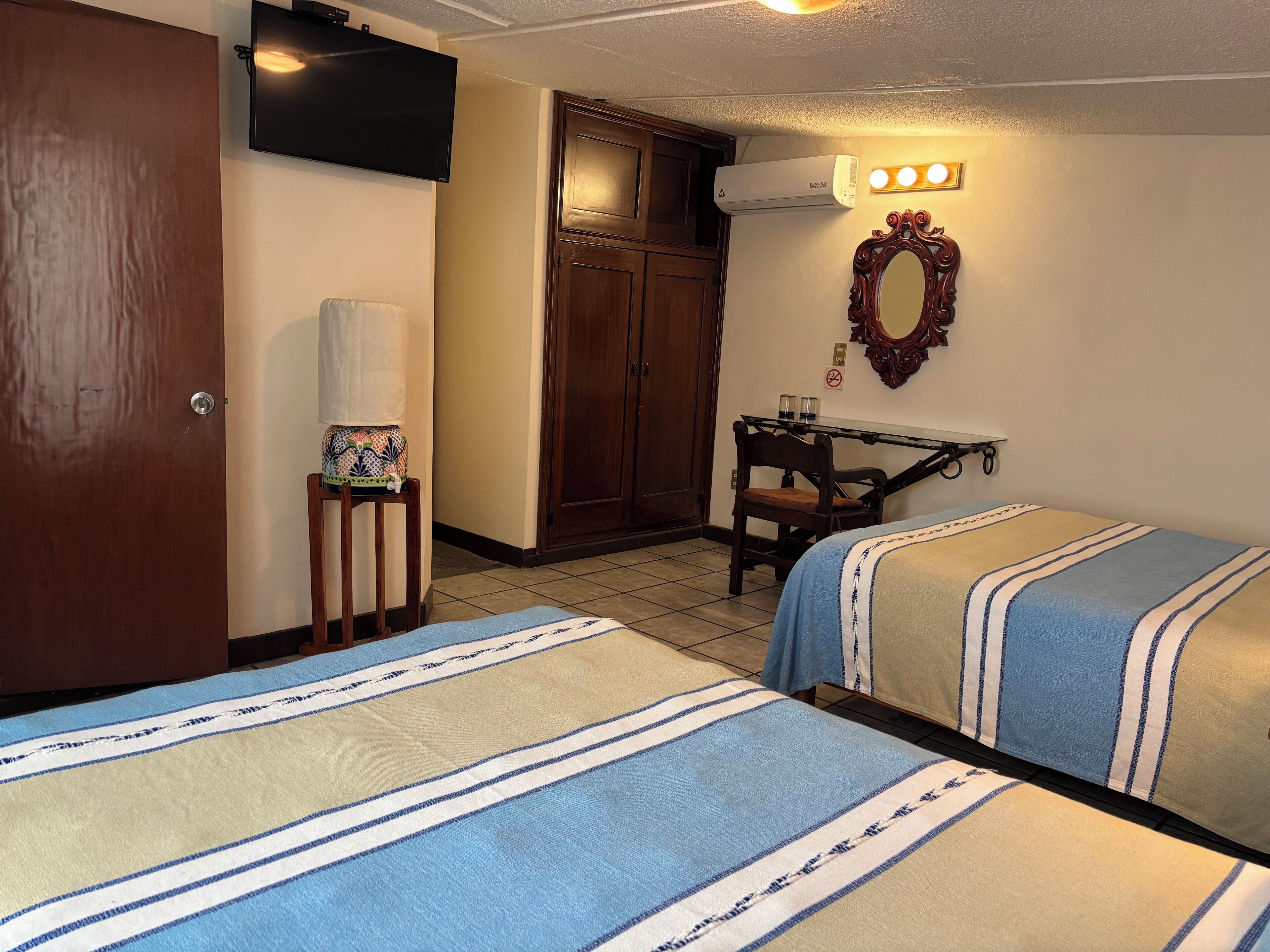 A hotel room with two beds, a desk with a chair, a TV, an air conditioner, and a mirror with lights.