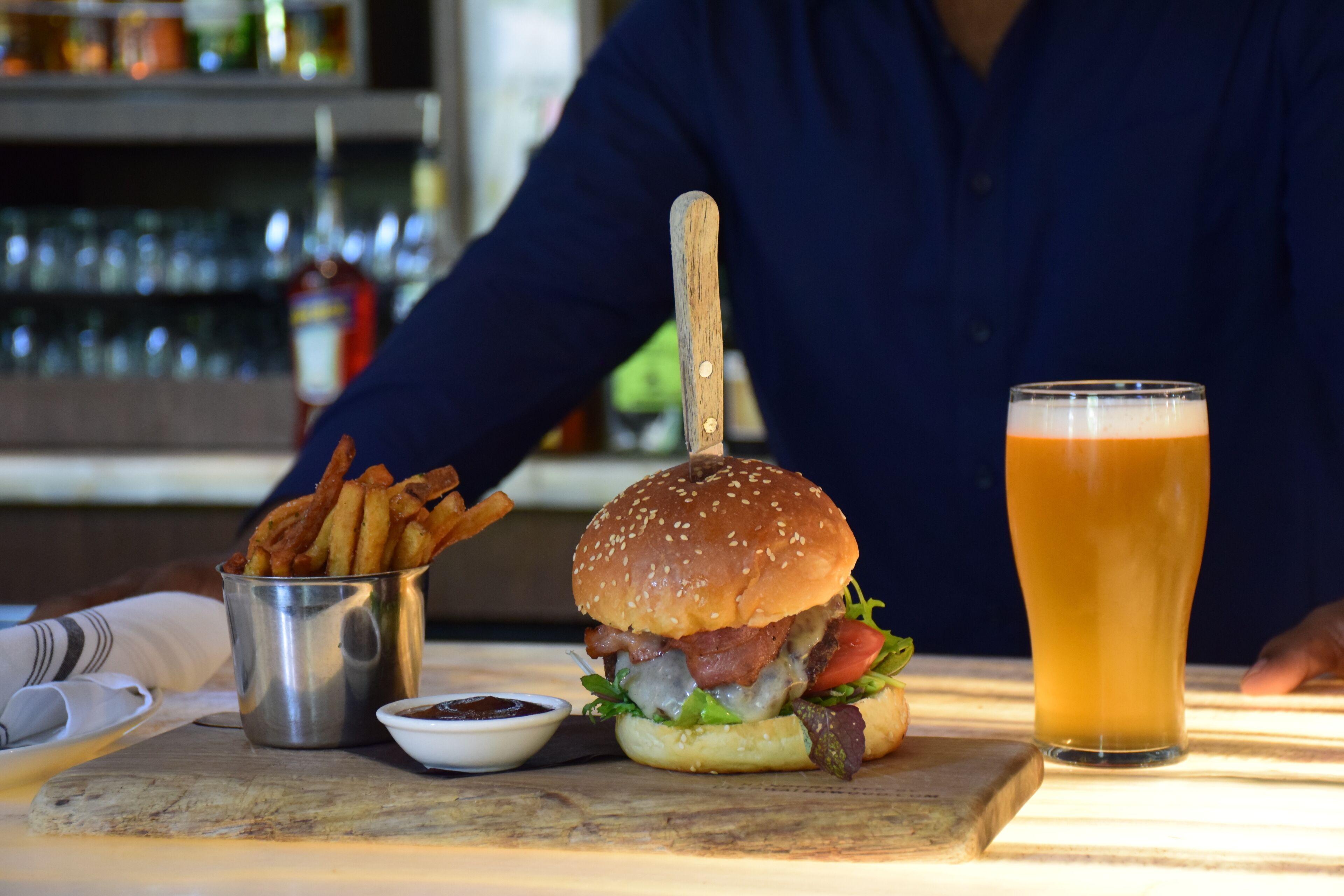 En burger med træspyd, et glas øl og en lille skål med sauce på et bord.