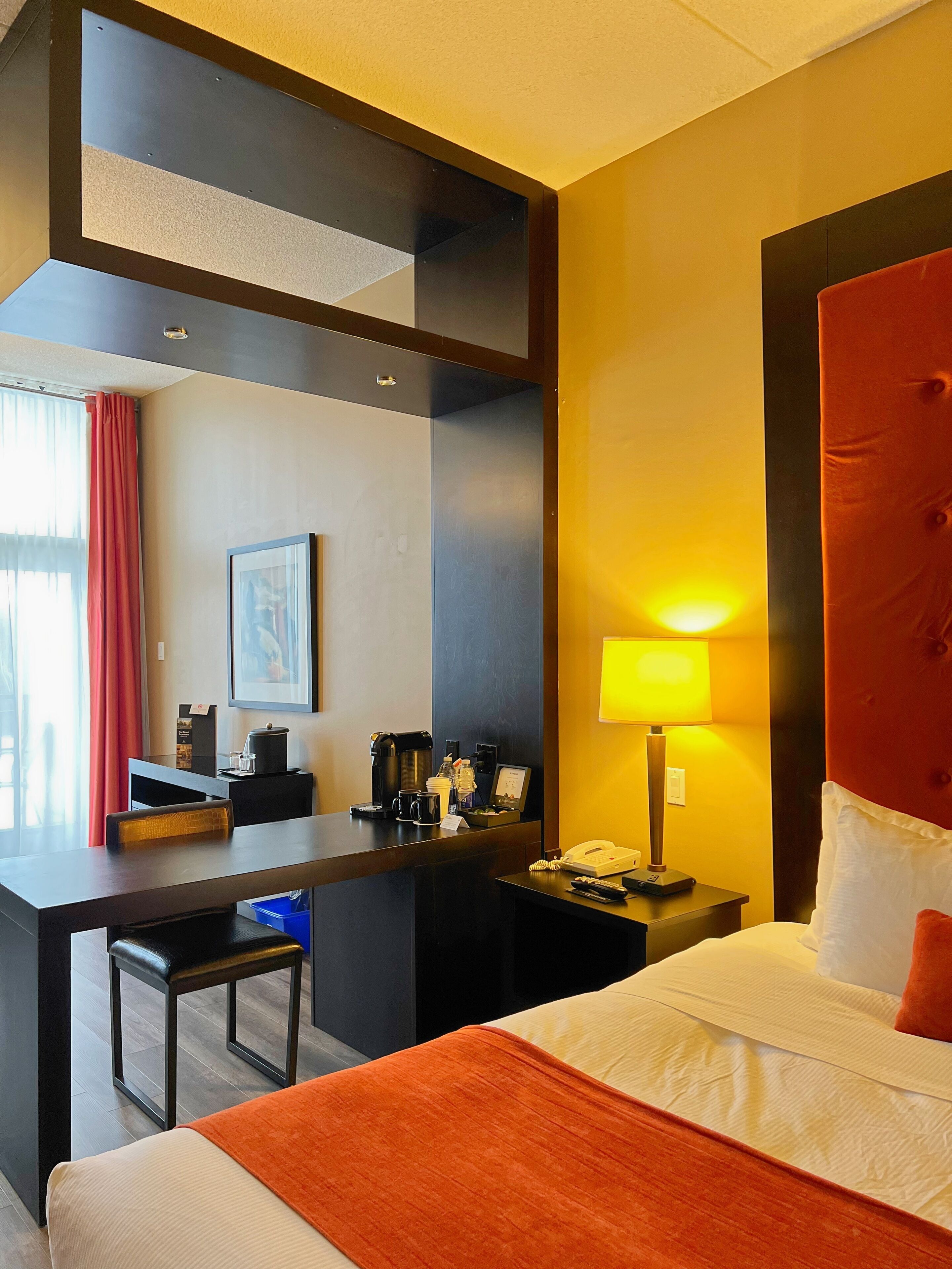 A hotel room with a bed, a desk, a chair, a lamp, a coffee machine, and a telephone.