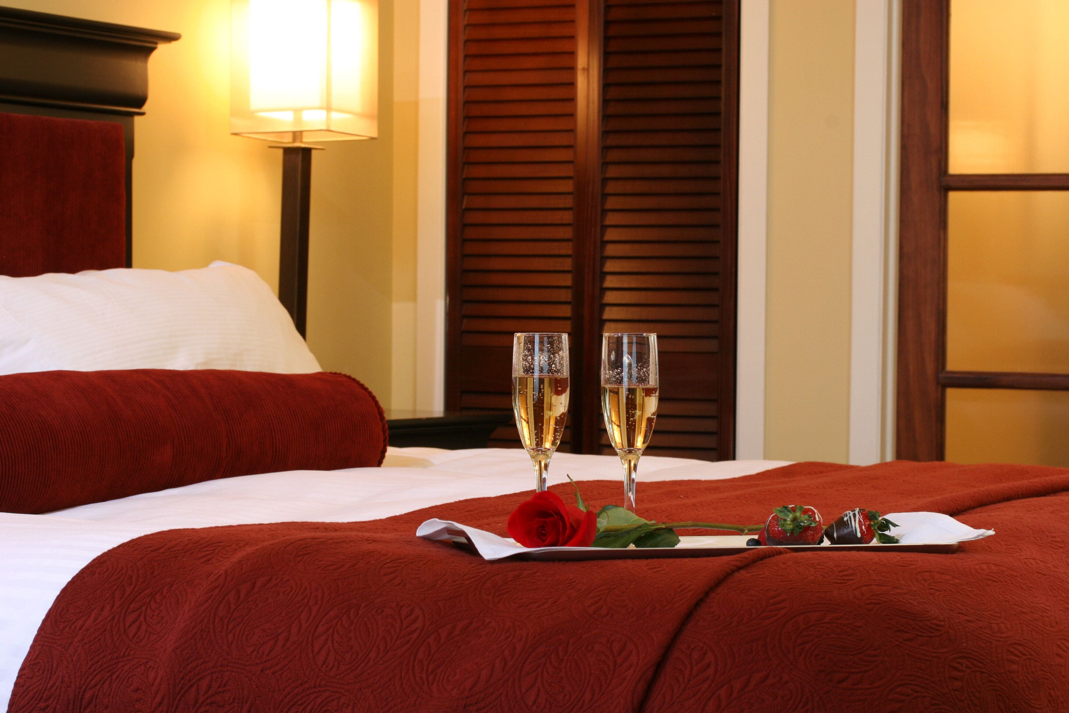 A neatly made bed with a red blanket, two champagne flutes, a rose, and strawberries on a tray.