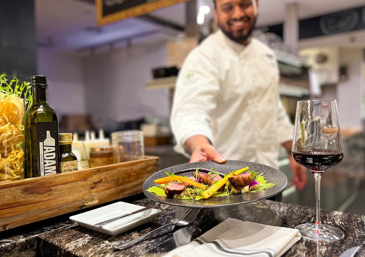 Un chef presenta un plato, con una botella de vino y una copa sobre la mesa.