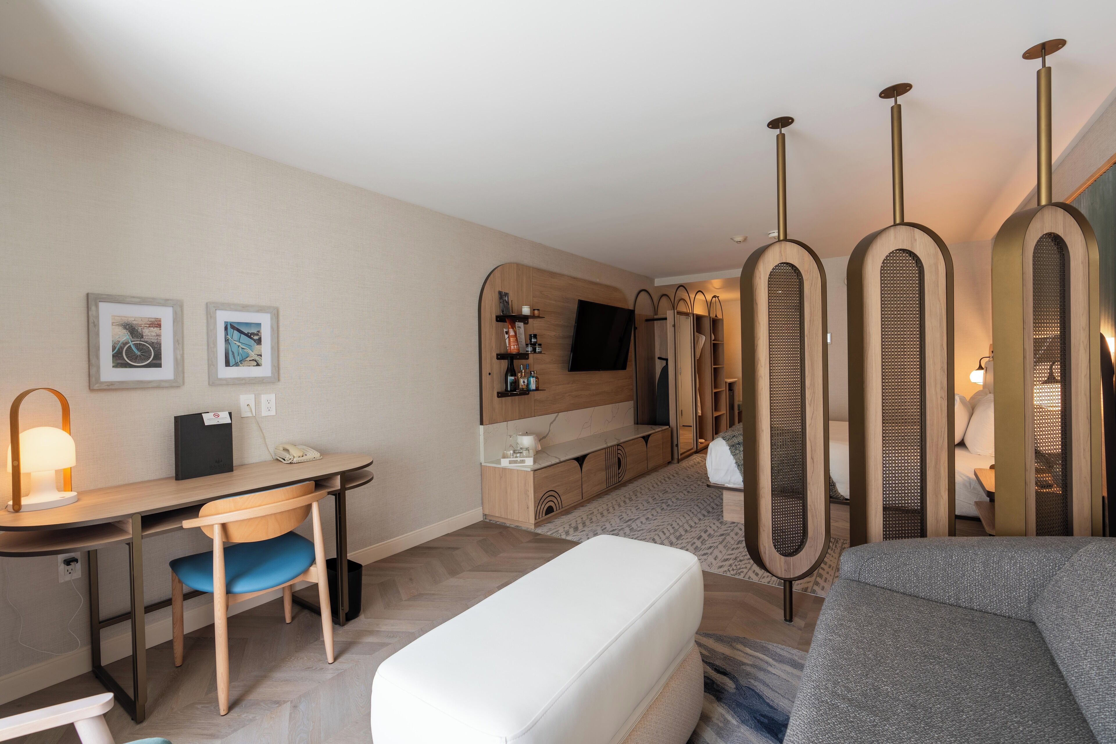 A modern hotel room with a wooden desk, a comfortable sofa, a flat-screen TV mounted on the wall, and a small seating area with a bench.