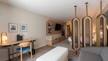 A modern hotel room with a wooden desk, a comfortable sofa, a flat-screen TV mounted on the wall, and a small seating area with a bench.