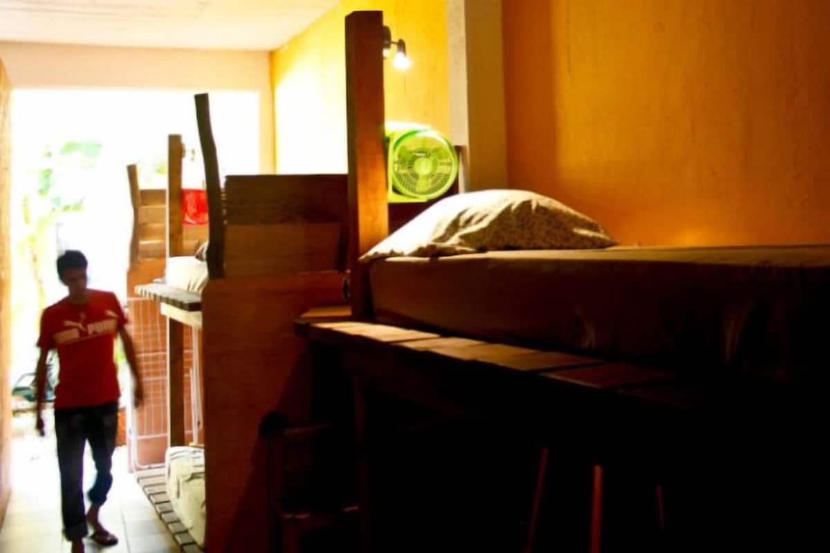 Une pièce faiblement  éclairée, avec un lit superposé, un ventilateur et une fenêtre.