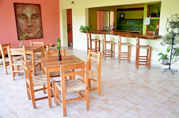 Un comedor con mesas y sillas de madera, una cocina al fondo y un cuadro grande en la pared.