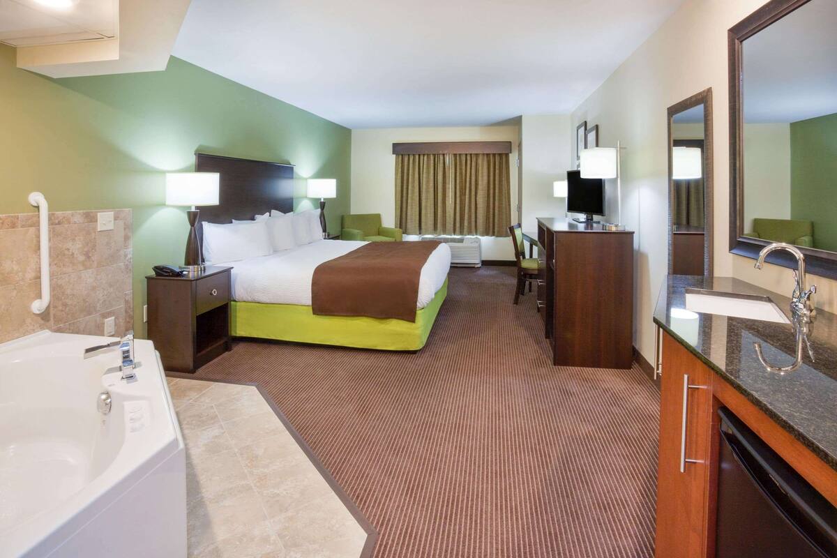Ein Hotelzimmer mit einem großen Bett, einem separaten Schreibtischbereich, einem Badezimmer mit Badewanne und einem großen Spiegel.