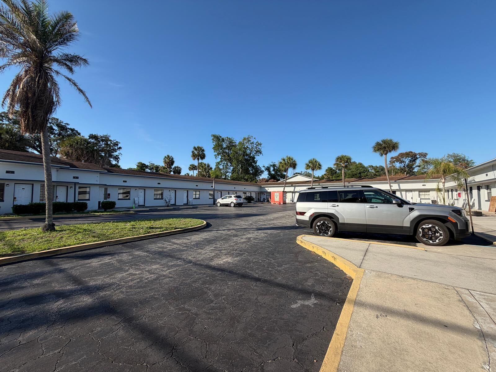A parking lot with a white SUV, a row of single-story buildings with white exteriors, and palm trees in the background.