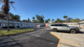 A parking lot with a white SUV, a row of single-story buildings with white exteriors, and palm trees in the background.
