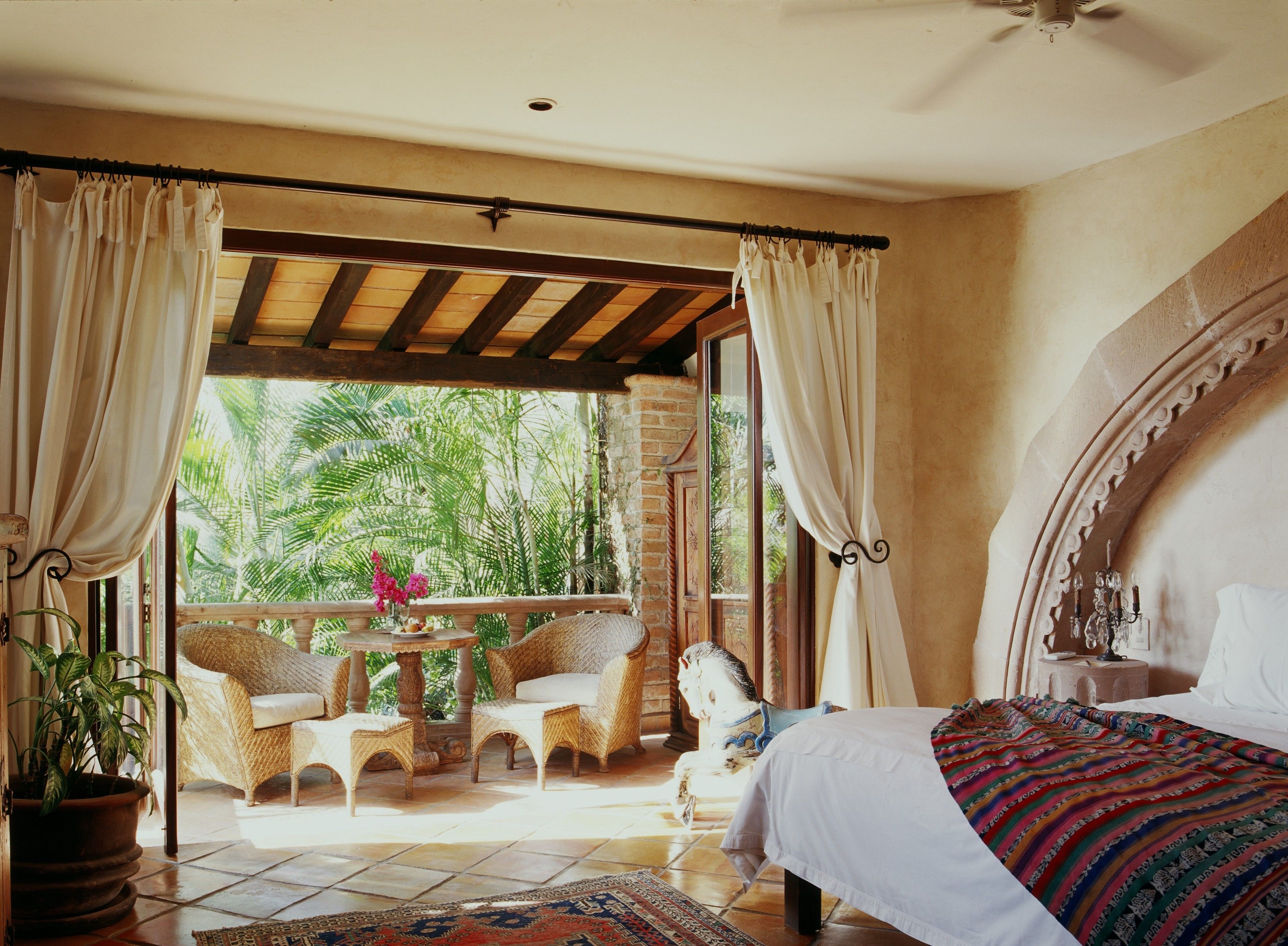 A room with a bed, a balcony with wicker furniture, and a view of tropical foliage.