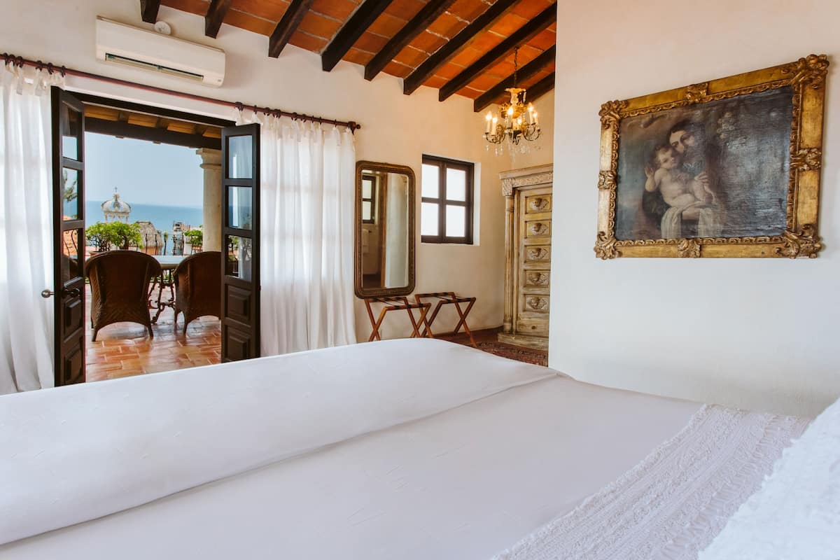 Een slaapkamer met een bed, een dressoir, een spiegel en een schilderij. Er is uitzicht op de zee door een geopende deur.
