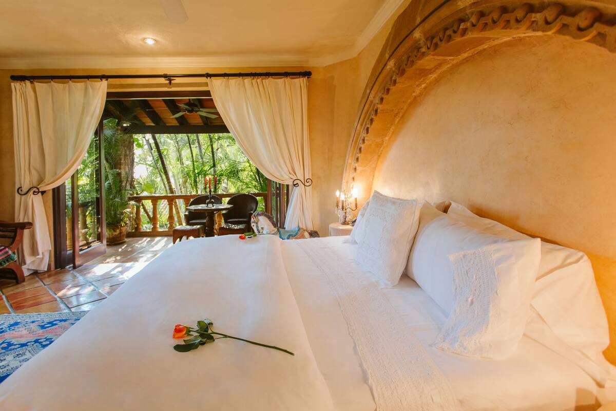 Een groot bed met witte lakens, een roos op het bed, en uitzicht op een buitenruimte met een tafel en stoelen.