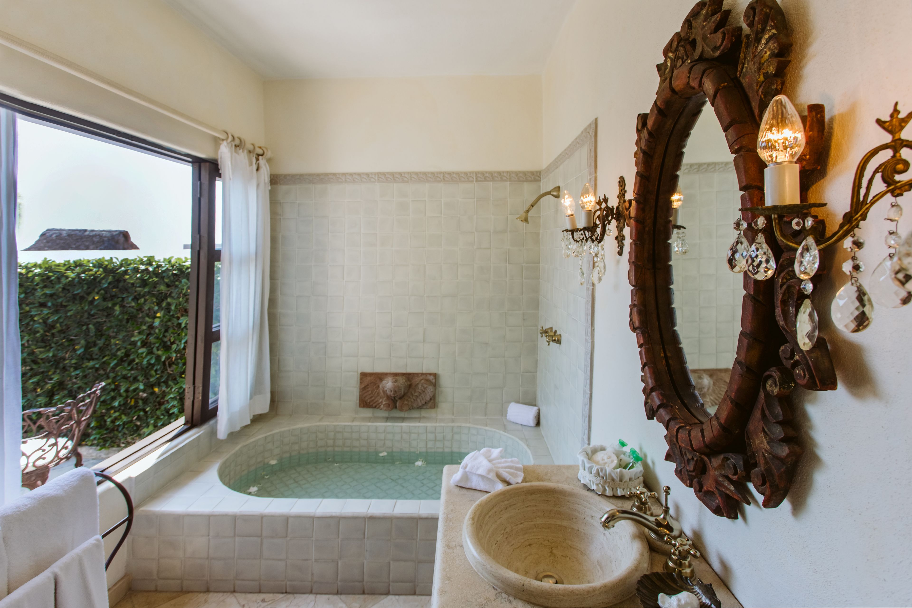 A bathroom with a large bathtub, a vanity with a bowl sink, a mirror with ornate decorations, and a window with curtains.
