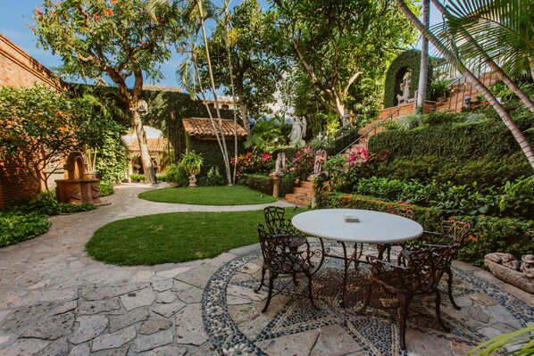 Une terrasse dotée d’une table et de chaises recouvertes de mosaïques, entourée d’une végétation luxuriante et bordée d’un mur de briques.