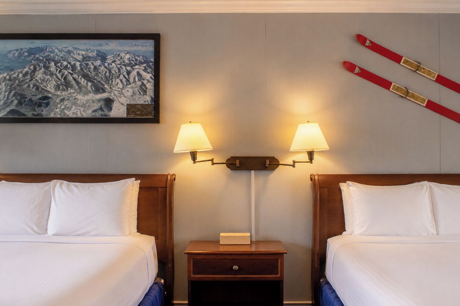 Habitación de hotel con dos camas, una mesita de noche con lámpara, un cuadro con paisaje de montaña y un par de esquíes en la pared.