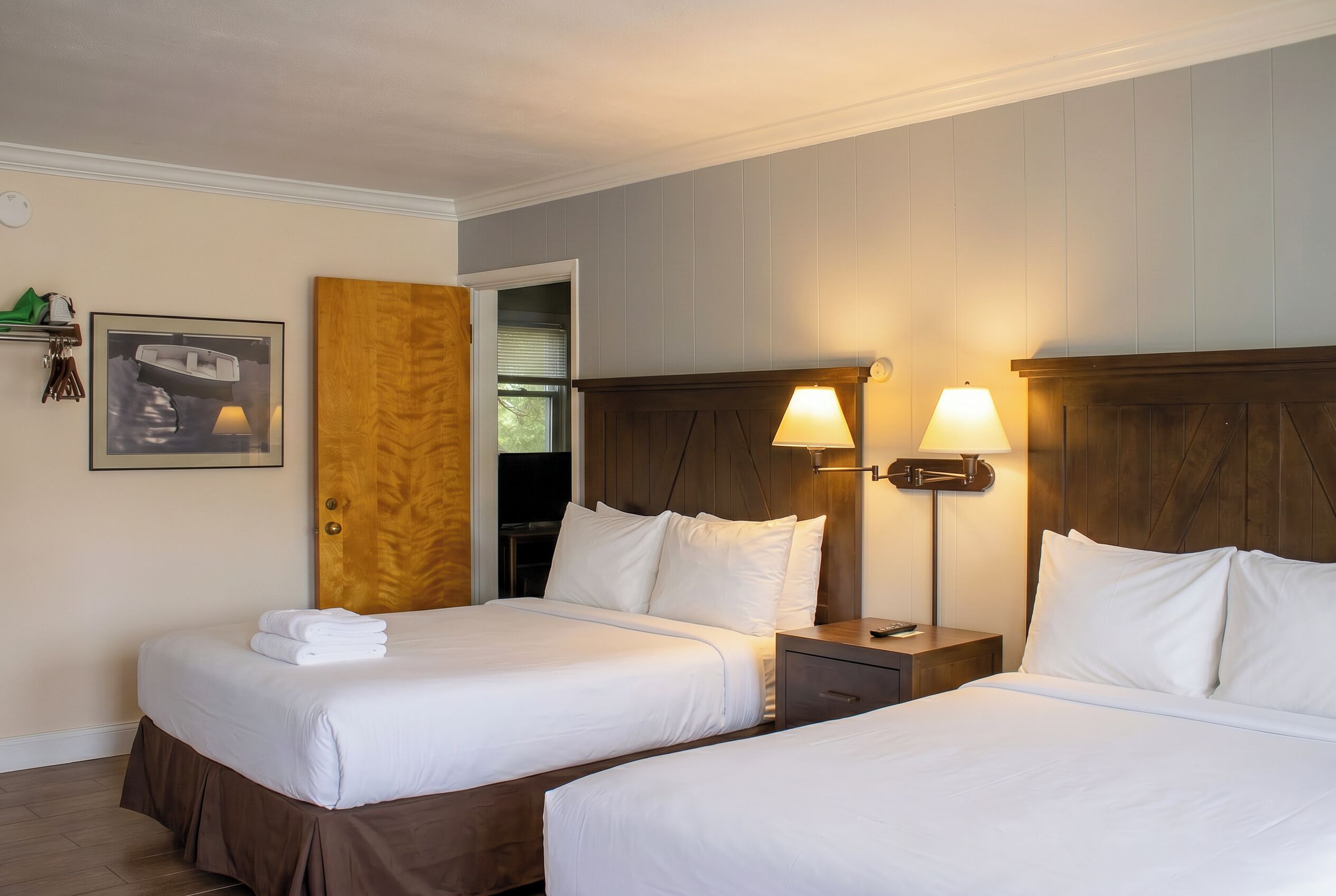 A hotel room with two beds, a wooden headboard, a nightstand with a lamp, a framed picture on the wall, and a door leading to another room.