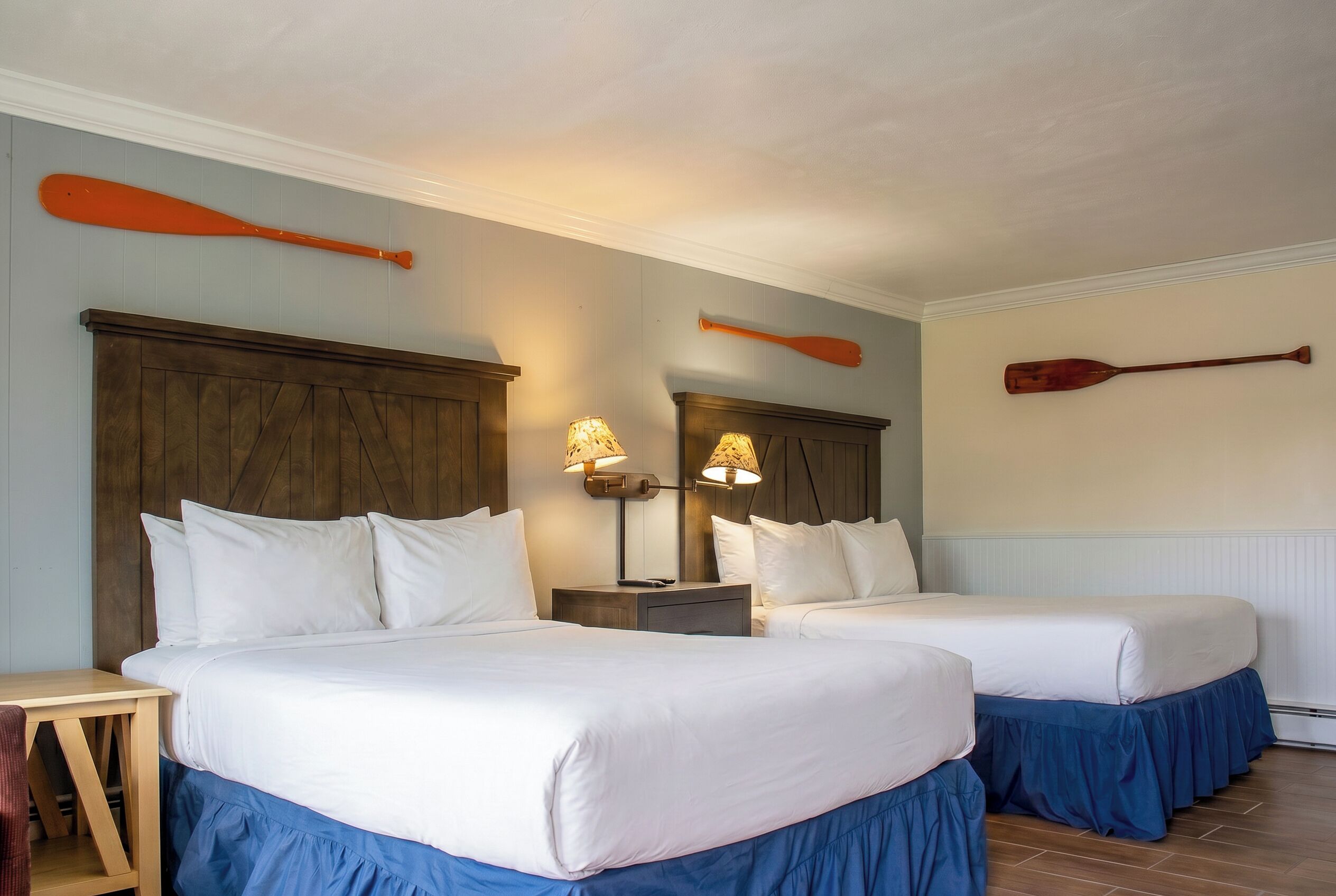 A hotel room with two beds, wooden headboards, a nightstand with lamps, and a paddle on the wall.