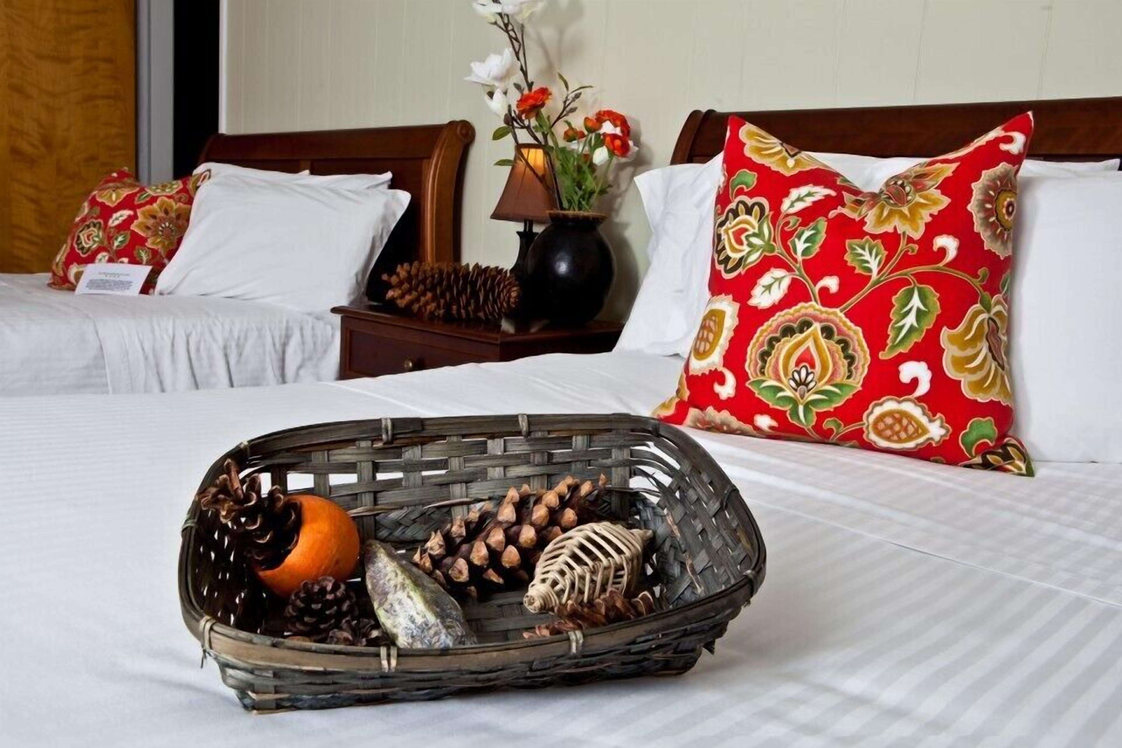 Habitación de hotel con dos camas, cada una con ropa de cama blanca y almohadas rojas con estampado floral. Una cesta de mimbre llena de piñas, una naranja y un objeto decorativo está sobre la cama.