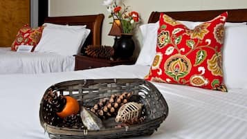 Habitación de hotel con dos camas, cada una con ropa de cama blanca y almohadas rojas con estampado floral. Una cesta de mimbre llena de piñas, una naranja y un objeto decorativo está sobre la cama.
