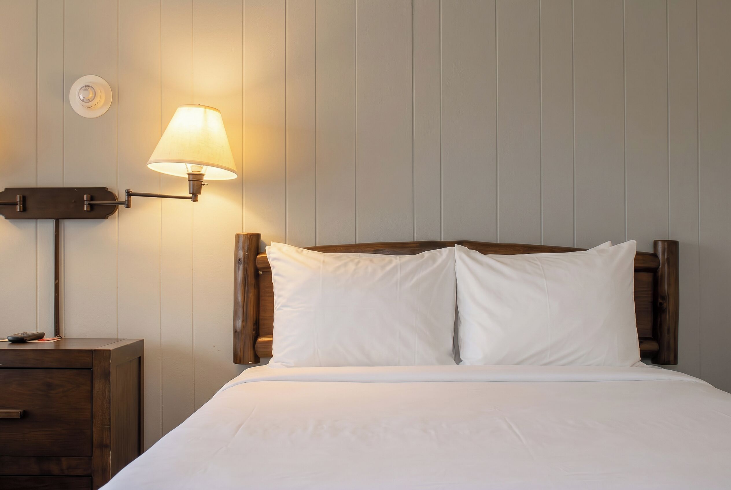 Una cama bien hecha con sábanas blancas, una mesita de noche de madera con una lámpara y una lámpara de pared.