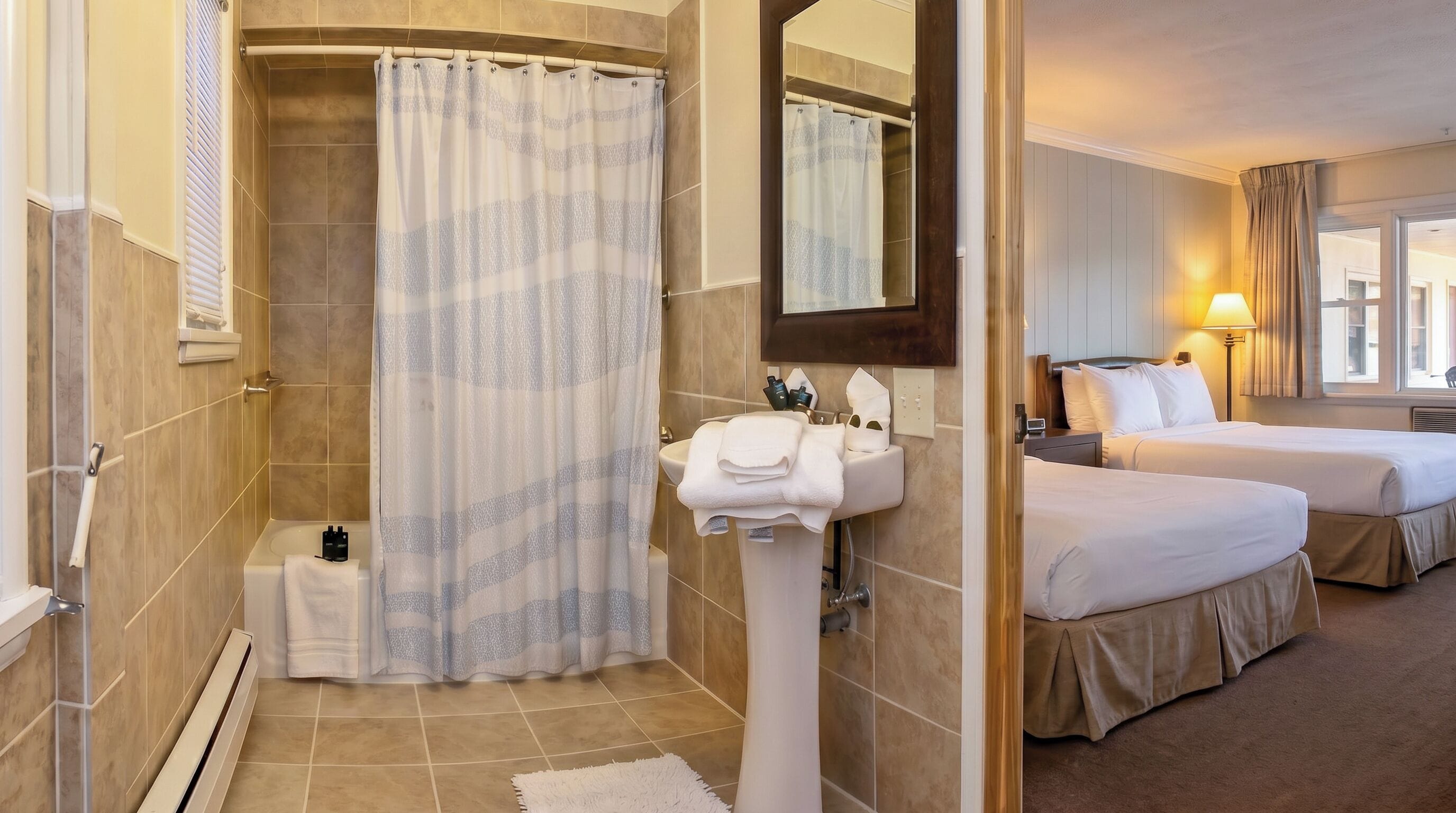 Une salle de bain d’hôtel avec une douche, un lavabo et un miroir. La pièce adjacente contient deux lits.
